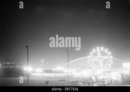 Foire la nuit., Whizgle News, Dutch Desk, pays-Bas, 1950 - 2000. Ce sont les éléments de l'image. La scène capture un carnaval nocturne animé, illuminé par un ensemble de lumières vives contre un ciel sombre. Une grande roue est bien en vue, son design complexe entouré de lumières scintillantes et vibrantes qui créent un effet étoilé. La roue domine les attractions environnantes, attirant le regard vers le haut. Au premier plan, divers stands et manèges de carnaval sont visibles, leurs structures projetant des ombres dramatiques sur le sol. Les tentes colorées et les cabines sont peu arran Banque D'Images