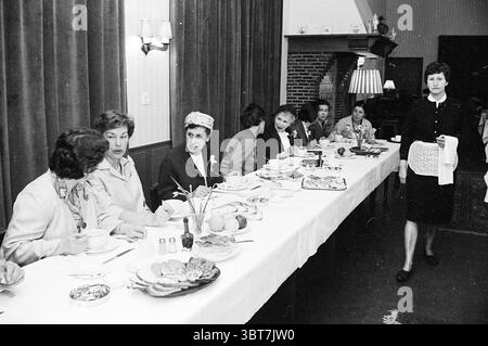 Les femmes au foyer irlandaises + les femmes de l'association des femmes au foyer d'IJmuiden déjeunent dans Velserend Groups, Whizgle News, Dutch Desk, pays-Bas, 1950 - 2000 le 04-05-1964. L'image montre ces rubriques. La scène capture un rassemblement à une longue table à manger dans un cadre intérieur. Des rideaux riches et sombres accrochent aux murs, créant une atmosphère chaleureuse et accueillante. Assis à la table sont plusieurs personnes engagées dans la conversation. Chaque personne affiche un style distinct, avec des vêtements allant des robes élégantes aux tenues plus décontractées, reflétant un mélange de la mode du milieu du XXe siècle. L'homme avec un n Banque D'Images