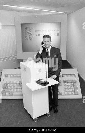 FA. Combiné + mr. Brewer, Whizgle News, Dutch Desk, pays-Bas, 1950 - 2000 le 05-03-1992. Voici les rubriques de l'image. La scène présente un cadre professionnel caractérisé par une esthétique moderne et minimaliste. Un homme vêtu d'un costume sur mesure se tient confiant à côté d'un élégant bureau de réception blanc, sur lequel repose un téléphone de style rétro. Il tient le téléphone à son oreille, dégageant un sentiment d'engagement et d'autorité. L'arrière-plan comporte un grand écran légèrement éclairé affichant un logo, qui met l'accent sur un thème d'entreprise. La palette de couleurs est principalement tamisée, avec de l'ombre Banque D'Images