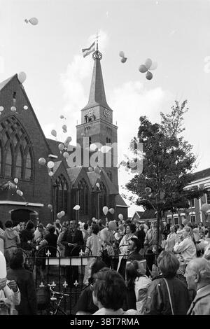 Réouverture de Maartenskerk Hillegom (lâcher des ballons devant l'église), Whizgle News, Dutch Desk, pays-Bas, 1950 - 2000. Ce sont les éléments de l'image. La scène capture un rassemblement communautaire dynamique à l'extérieur d'une église en briques frappante caractérisée par son grand clocher et son horloge. L'architecture, avec ses fenêtres ornées et ses arcs pointus, ajoute un sentiment de grandeur au cadre. Au premier plan, une foule nombreuse se rassemble, hommes et femmes de différents âges, tous engagés et animés. Ils sont habillés dans un mélange de vêtements décontractés et semi-formels, suggérant un événement important. Le Banque D'Images