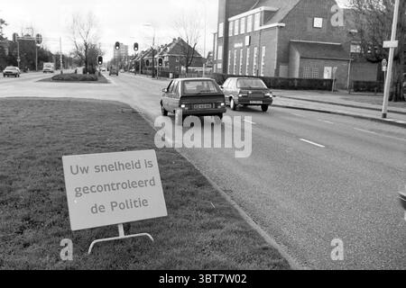 Speed control Heemsteedse Dreef control Heemstede Heemsteedse Dreef pays-Bas, Whizgle News, Dutch Desk, pays-Bas, 1950 - 2000 on 11-01-1988. L'image inclut ces rubriques. La scène représente une zone de circulation le long d'une route achalandée, avec deux voitures se déplaçant dans des directions opposées. Les véhicules sont différents modèles, dont l'un semble légèrement plus ancien, et les deux sont principalement dans des tons sombres comme le bleu et le noir. À droite, il y a une étendue d'herbe bordée par un trottoir en béton, offrant une séparation claire de la route. Un grand panneau se dresse sur l'herbe, sa structure audacieuse dessine Banque D'Images