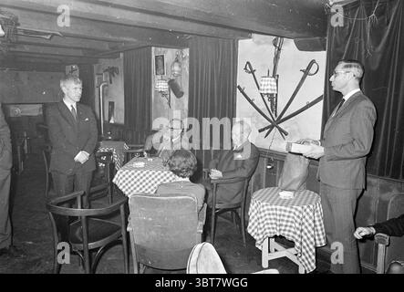 Présentation de GIFT, Whizgle News, Dutch Desk, pays-Bas, 1950 - 2000 le 03-1969. L'image inclut ces rubriques. La scène se déroule dans une pièce confortable et faiblement éclairée qui dégage un charme vintage, caractérisé par des poutres en bois sombres à travers le plafond. Les murs sont ornés d'un mélange de décor rustique et de textures subtiles, contribuant à une atmosphère accueillante et intime. Au premier plan, un groupe d'individus bien habillés s'engage dans la conversation. Une personne, debout à gauche, porte un costume sur mesure et semble parler, peut-être prononcer un discours ou faire un point. Il l'a fait Banque D'Images