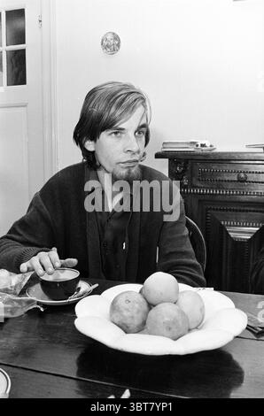 Photo de groupe et portraits de 4 jeunes hommes., Whizgle News, Dutch Desk, pays-Bas, 1950 - 2000. Ces rubriques sont illustrées dans l'image. Dans une salle à manger chaleureusement éclairée, un jeune homme est assis à une table en bois, occupant le centre de la scène. Son expression réfléchie suggère la contemplation, avec un soupçon d'intrigue dans ses yeux. Il porte un cardigan de couleur foncée, et ses cheveux soignés encadrent son visage, accentuant ses traits faciaux, y compris une barbe claire qui ajoute à son apparence mature mais jeune. Sur la table devant lui se trouve un grand plateau rond rempli de plusieurs oranges, leur vi Banque D'Images