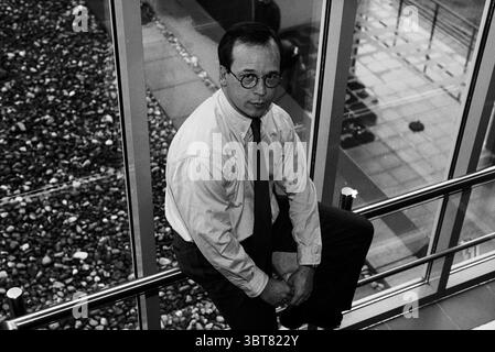 Mr. Sack Ditmar Boekenimport], Whizgle News, Dutch Desk, pays-Bas, 1950 - 2000 on 31-10-1996. Ces rubriques apparaissent dans l'image. Dans cette scène, un homme s'assoit sur une rampe, positionnée légèrement au-dessus de ce qui semble être un espace moderne et ouvert. Il est vêtu d'une chemise boutonnée de couleur claire et porte des lunettes rondes, lui donnant une apparence intellectuelle. Sa cravate foncée contraste fortement avec la chemise plus légère. L'arrière-plan présente un grand mur de verre, permettant à la lumière naturelle de filtrer, créant une atmosphère lumineuse et aérée. Juste à l'extérieur du verre, il y a un lit de STO lisse et rond Banque D'Images