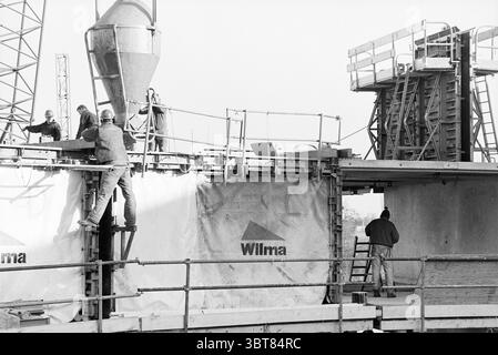 Construction de maisons C42 H'lem Haarlem pays-Bas, Whizgle News, Dutch Desk, pays-Bas, 1950 - 2000 le 01-02-1995. Voici les rubriques de l'image. La scène capture un chantier de construction actif, animé par l'activité. Au premier plan, les travailleurs sont engagés dans diverses tâches, leurs postures indiquant la concentration et le travail d'équipe. Un individu se tient au sommet d'un échafaudage, se préparant à guider une grande goulotte d'où un flux de béton émergera, mettant en valeur l'équipement lourd typique de tels environnements. Les travailleurs sont habillés de vêtements fonctionnels, principalement avec des nuances Banque D'Images