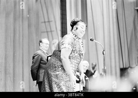 Soirée NZH Noord Zuid Hollandse Vervoersmaatschappij N.V. NZ, Whizgle News, Dutch Desk, pays-Bas, 1950 - 2000 on 10-10-1970. Ces rubriques sont illustrées dans l'image. La scène représente une performance ou un événement dynamique, caractérisé par une énergie et un engagement vifs. Au premier plan, une femme se tient devant un micro, son expression animée et joyeuse, suggérant un moment de connexion avec son public. Elle porte une robe richement texturée qui présente des motifs élaborés, probablement dans des tons de couleurs profondes comme Bordeaux ou émeraude, renforçant sa présence sur scène. À gauche, un homme est visibl Banque D'Images