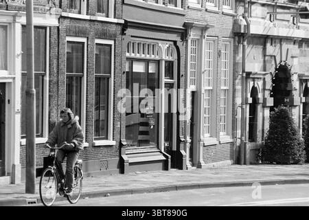 Antiquaires Damstraat Haarlem Damstraat pays-Bas, Whizgle News, Dutch Desk, pays-Bas, 1950 - 2000 le 06-10-1994. Ces rubriques apparaissent dans l'image. La scène se déroule dans un environnement urbain caractérisé par une rangée de charmants bâtiments anciens présentant une architecture traditionnelle. Les façades sont composées de différentes textures, avec des détails en brique et en pierre, et de grandes fenêtres qui reflètent la lumière silencieuse du jour. Un cycliste occupe le premier plan, conduisant un vélo noir classique. Ils sont habillés d'un manteau de couleur claire avec une capuche doublée de fourrure, suggérant un climat frais. Le cycliste Banque D'Images