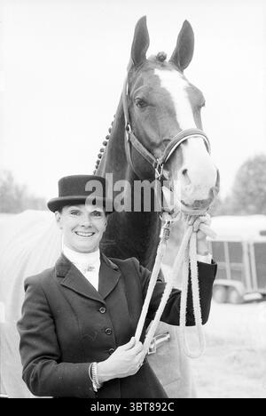 Concours de chevaux., Whizgle News, Dutch Desk, pays-Bas, 1950 - 2000. Ces rubriques sont illustrées dans l'image. Dans cette scène, une femme se tient fièrement à côté d'un grand cheval. La femme respire la confiance et la joie, portant un élégant manteau noir qui contraste magnifiquement avec le manteau brillant du cheval. Son élégant chapeau haut de gamme ajoute une touche de sophistication, tandis que son sourire éclatant souligne son lien avec l'animal. Le cheval, dominant à côté d'elle, a un manteau frappant avec une riche couleur de baie et un flambeau blanc proéminent qui court sur son visage. Sa crinière bien entretenue est soigneusement bandelée, suggérant des soins et Banque D'Images