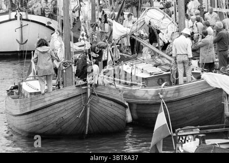 Les gens sur leur bateau dans le port pendant les festivités, Whizgle News, Dutch Desk, pays-Bas, 1950 - 2000 en 1990. L'image montre ces rubriques. La scène présente un port animé rempli de bateaux en bois, leurs surfaces lisses reflétant les mouvements subtils de l'eau. Deux vaisseaux proéminents, gracieusement courbés dans la forme, sont ancrés près l'un de l'autre, mettant en valeur le savoir-faire complexe dans leur conception. Des gens d'âges et de styles variés sont dispersés dans les bateaux et le long du rivage, certains engagés dans la conversation tandis que d'autres semblent être occupés par les préparatifs, peut-être pour une veille Banque D'Images