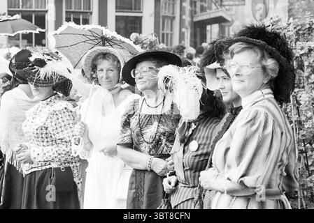 Dames en vêtements historiques devant la mairie, Whizgle News, Dutch Desk, pays-Bas, 1950 - 2000 le 31-08-1979. L'image montre ces rubriques. La scène capture un groupe de femmes vêtues d'une tenue élégante d'une époque passée, chacune ornée de chapeaux au design complexe, certains ornés de plumes et de fleurs. Les femmes se tiennent étroitement ensemble, démontrant un sentiment de camaraderie. Leurs tenues reflètent un mélange de textures et de motifs : des motifs floraux aux rayures, mettant en valeur les divers choix de mode de l'époque. L'arrière-plan présente une scène de rue pittoresque, avec quelques structures Banque D'Images