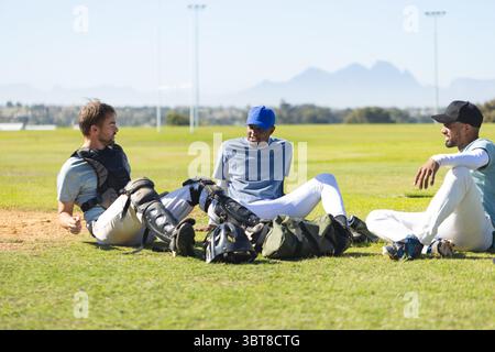 Divers coéquipiers masculins assis et discutant pendant la pause sur le terrain de baseball avec des casques et des gants Banque D'Images