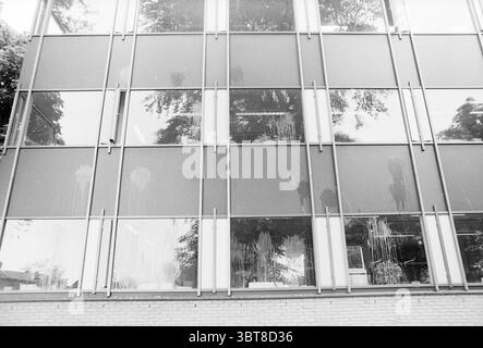 Hôtel de ville teinté d'oeufs Hillegom vandalisme Hillegom, Whizgle News, Dutch Desk, pays-Bas, 1950 - 2000 on 16-07-1986. Ces rubriques apparaissent dans l'image. La scène présente un bâtiment moderne caractérisé par une grande façade en verre, qui reflète son environnement. Les panneaux de verre semblent striés et partiellement obscurcis, suggérant qu'ils ont été éclaboussés d'une substance inconnue, peut-être de la peinture. Cela donne un sentiment de négligence ou de délabrement. En arrière-plan, les arbres sont visibles, leurs branches projetant des ombres complexes sur la surface du verre, créant un jeu de lumière et d'obscurité. Le buildi Banque D'Images