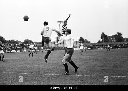 Football Haarlem pays-Bas, Whizgle News, Dutch Desk, pays-Bas, 1950 - 2000 en 1964. Ce sont les éléments de l'image. La scène capture un moment d'excitation sur un terrain de sport herbeux, avec une atmosphère animée autour d'un match de football. Trois joueurs sont en vedette : un dans un kit blanc avec le numéro 9, sautant dans les airs dans une pose dynamique, tandis que deux autres dans des shorts rayés et sombres distincts sont engagés dans le jeu. Le joueur en blanc semble tenter de prendre le contrôle d'une balle qui est suspendue dans les airs. Le fond est rempli de spectateurs, suggérer Banque D'Images