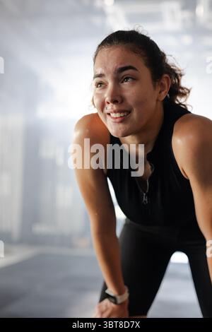 Athlète féminine en sportswear noir appuyé sur les genoux respirant fort dans la salle de gym portant une smartwatch blanche Banque D'Images
