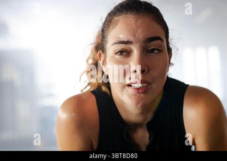 Athlète féminine penchée en avant après l'entraînement parmi les machines de gym floues, portant un débardeur noir Banque D'Images