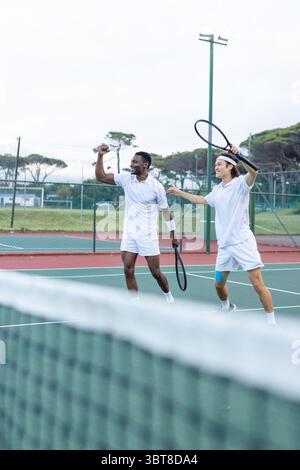Divers amis masculins célébrant le point de victoire tout en marchant sur un court de tennis extérieur avec des raquettes Banque D'Images