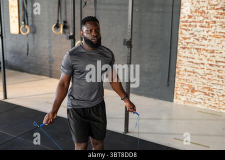 Homme afro-américain à sauter corde avec poignées bleues sur tapis dans un studio de fitness avec anneaux de gymnastique Banque D'Images