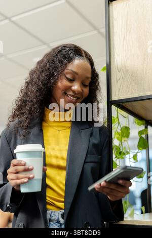 Femme afro-américaine tenant un smartphone défilant de tasse de café bleu dans la salle de pause à côté de l'étagère à plantes Banque D'Images