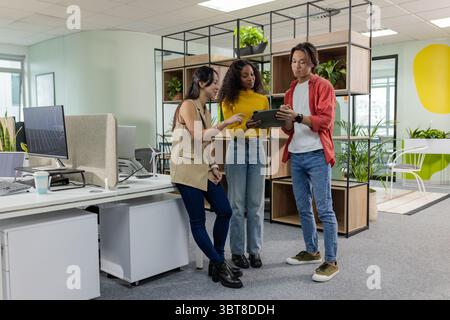 Divers collègues debout près d'étagères remplies de plantes discutant du projet sur tablette dans un bureau moderne Banque D'Images