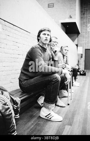 Portrait entraîneur de football en salle partenaire du maillot féminin : TEN Cate BV Agence de distribution Alkmaar, Whizgle News, Dutch Desk, pays-Bas, 1950 - 2000 sur 10-1980. Ces rubriques sont illustrées dans l'image. Dans un cadre intérieur spacieux ressemblant à une salle de sport ou à une salle de sport, plusieurs personnes sont assises sur un long banc en bois. Le sol a une finition en bois poli, améliorant l'atmosphère chaleureuse et accueillante. Les murs de briques offrent une toile de fond rustique, contrastant avec les éléments plus modernes. La personne au centre a une expression contemplative, assise de façon désinvolte avec les jambes croisées. Il porte une texture douillette Banque D'Images