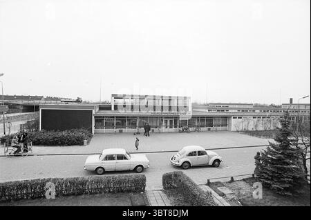 Haarlemmermeerlyceum, Whizgle News, Dutch Desk, pays-Bas, 1950 - 2000 on 01-01-1973. L'image montre ces rubriques. La scène capture un cadre architectural du milieu du siècle caractérisé par une vue extérieure large et ouverte d'un bâtiment. La structure a un design moderniste avec un toit plat et de grandes fenêtres qui reflètent une esthétique utilitaire. Au premier plan, deux voitures classiques sont garées le long d'une zone pavée ; l'une est une nuance de gris clair tandis que l'autre est un blanc doux, les deux ajoutant une touche vintage à l'atmosphère. Quelques personnes peuvent être vues interagir dans l'espace ; une figure est le purpo marchant Banque D'Images