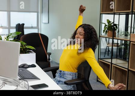 Femme afro-américaine s'étirant au bureau avec des étagères de plantes de smartphone d'ordinateur Banque D'Images