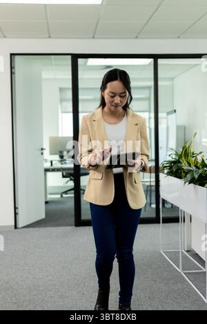Femme asiatique marchant dans le couloir du bureau devant les jardinières tapant sur la tablette portant blazer Banque D'Images
