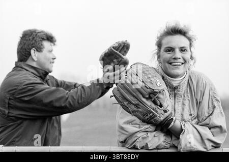 Natalie Kroonsberg & Johan Kluyskens, Whizgle News, Dutch Desk, pays-Bas, 1950 - 2000 le 14-04-1993. L'image contient ces rubriques. Dans un cadre extérieur dynamique, une scène vibrante se déroule sur un ciel silencieux. Une femme se tient bien en vue au premier plan, son sourire rayonnant de chaleur et de confiance. Elle porte un coupe-vent de couleur claire, et ses cheveux ondulés encadrent son visage, ajoutant à son comportement joyeux. Elle tient un gant de baseball d'une main, le posant désinvolte sur une balustrade. Derrière elle, un homme peut être vu, partiellement détourné, engagé dans son propre moment alors qu'il se prépare à lancer une baseba Banque D'Images