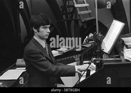 Organiste Frans Geels Orgues et organistes, Whizgle News, Dutch Desk, pays-Bas, 1950 - 2000 le 01-06-1970. Ces rubriques sont illustrées dans l'image. Dans un cadre intérieur légèrement éclairé, l’accent est mis sur un jeune musicien assis devant un piano à queue. Il a les cheveux foncés et est habillé dans un costume sur mesure qui transmet un sens de formalité. L'expression sur son visage est concentrée mais contemplative, reflétant son engagement avec la musique. Autour de lui se trouve un tableau de feuilles de musique, certaines soigneusement organisées et d'autres légèrement désalignées, indiquant une utilisation active. La surface polie du piano reflète subtilement les sons Banque D'Images