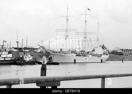 Navire d'entraînement Pollux amarré à IJmuiden navires IJmuiden pays-Bas, Whizgle News, Dutch Desk, pays-Bas, 1950 - 2000 le 10-01-1989. Ce sont les éléments de l'image. La scène présente un grand voilier, caractérisé par ses trois hauts mâts, chacun truqué de lignes et de voiles qui lui donnent une apparence majestueuse et vintage. La coque du navire est peinte en blanc, se distinguant par les tons sourds de l'environnement environnant. Le cadre est un port, où le navire est ancré aux côtés de plus petits navires, probablement des remorqueurs, indiqués par leur conception plus industrielle. Le contexte i Banque D'Images