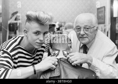 Coiffeur jeune et vieux avec tondeuses à cheveux Nassaustraat coiffeurs et salons de coiffure Haarlem Nassaustraat pays-Bas, Whizgle News, Dutch Desk, pays-Bas, 1950 - 2000 on 20-11-1987. L'image contient ces rubriques. Dans un salon de coiffure animé, deux figures sont au centre de l'attention. À gauche, une jeune personne avec une coiffure audacieuse qui présente un haut volumineux et stylé et des côtés rasés porte une chemise rayée noire et blanche. Leur expression est confiante, affichant un soupçon de sourire aux coins de leur bouche alors qu'ils tiennent des tondeuses contre l'arrière du cou d'un client, indiquant qu'ils sont Banque D'Images