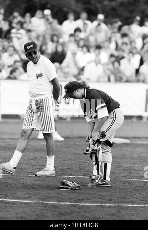 Softball Chinese T. - Sharks [Taiwan - Whiteford Sharks (USA)], Whizgle News, Dutch Desk, pays-Bas, 1950 - 2000 le 25-07-1991. Voici les rubriques de l'image. Dans un environnement sportif animé en plein air, la scène capture un moment pendant un match de baseball. Le champ est d'un brun riche, sa surface légèrement poussiéreuse, entouré d'un terrain vert vif. Se prélassant sur la touche, une foule de spectateurs est visible en arrière-plan, leurs expressions étant un mélange d’engagement et d’amusement. Au premier plan, il y a deux individus sur le terrain. Une personne, un homme avec une posture joviale, porte un col léger Banque D'Images