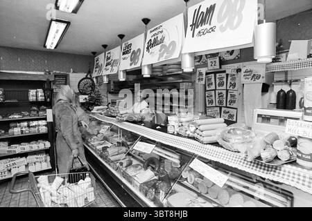 Intérieur d'un supermarché., Whizgle News, Dutch Desk, pays-Bas, 1950 - 2000. Voici les rubriques de l'image. La scène capture un décor traditionnel de charcuterie ou d'épicerie, imprégné d'une esthétique vintage en noir et blanc. Une femme se tient debout au comptoir, vêtue d'un long manteau, la tête partiellement couverte, suggérant une atmosphère fraîche. Elle semble engager une conversation avec le membre du personnel derrière le comptoir, qui porte un tablier et présente un comportement invitant. Le comptoir présente une gamme de charcuteries, fromages et divers produits emballés, tous soigneusement disposés dans un verre di Banque D'Images