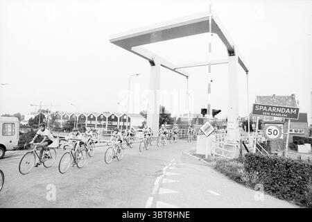 Ronde de moustiques au pont Spaarndam Muggeronde, Whizgle News, Dutch Desk, pays-Bas, 1950 - 2000 le 27-07-1986. Ce sont les éléments de l'image. Dans cette scène, un groupe de cyclistes s'approche d'une grande structure de pont moderne qui vole au-dessus. Les cyclistes portent des casques et des maillots assortis, mettant l'accent sur un esprit sportif ; leurs vélos sont élégants et conçus pour la vitesse. Le cadre semble être une zone de banlieue, avec un mélange de bâtiments résidentiels en arrière-plan présentant un style architectural minimaliste avec des toits en pente et une palette de couleurs sourdes. La route est une double CA Banque D'Images
