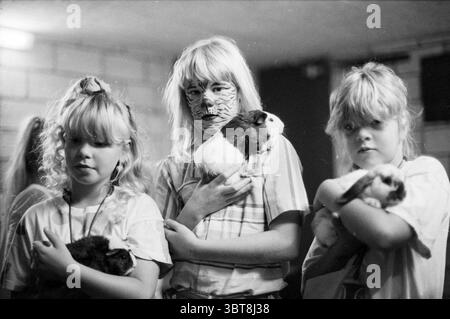 Poussière poussière enfant et poussière animale, Whizgle News, Dutch Desk, pays-Bas, 1950 - 2000 le 09-08-1989. L'image contient ces rubriques. Dans un espace intérieur légèrement éclairé, trois enfants se tiennent proches les uns des autres, berçant chacun un petit cobaye. Le décor présente des murs de briques apparentes qui contribuent à une atmosphère rustique et légèrement tamisée. Les enfants, tous avec des cheveux de couleur claire, affichent des coiffures variées : l'un a des queues de cochon, tandis qu'un autre arbore un look texturé et moelleux. La jeune fille au centre a son visage orné de rayures saisissantes ressemblant à un tigre dans la peinture sombre, ce qui ajoute un élément de ludique Banque D'Images