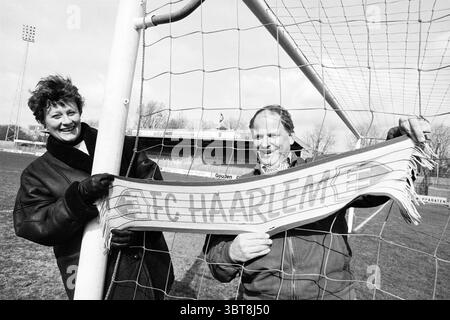 Famille Wiersma avec FC Haarlem Scarf, Whizgle News, Dutch Desk, The Netherlands, 1950 - 2000 le 24-03-1995. L'image inclut ces rubriques. Dans un cadre sportif et extérieur, deux individus se tiennent ensemble près du poteau d’un terrain de football, leurs visages étant éclairés par la lumière naturelle. La toile de fond présente un stade animé, montrant sa structure et des tribunes lointaines remplies de spectateurs. La première personne, une femme, dégage un comportement joyeux. Ses cheveux courts et touffus suggèrent une journée venteuse, et son sourire transmet l'enthousiasme. Elle porte une veste sombre qui contraste avec la scène vibrante autour d'elle. Suivant Banque D'Images