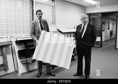 Hangar avec matériaux de construction, Whizgle News, Dutch Desk, pays-Bas, 1950 - 2000 on 21-12-1982. Voici les rubriques de l'image. Dans la scène, deux hommes se tiennent en évidence dans un cadre intérieur caractérisé par une esthétique moderne du milieu du siècle. Un homme, portant un costume de couleur claire avec une cravate et des cheveux soigneusement peignés, tient une grande feuille de matériau, mettant en valeur sa texture ondulée. L’autre homme, vêtu d’un costume plus sombre avec une cravate, se tient à côté de lui, son expression véhiculant un mélange d’autorité et de curiosité. L'arrière-plan présente un environnement de bureau spacieux, avec des rangées d'étalages Banque D'Images
