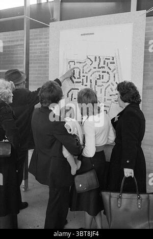 Présentation des plans de construction (maison), Whizgle News, Dutch Desk, pays-Bas, 1950 - 2000 en 1977. Ces rubriques apparaissent dans l'image. Dans la scène, un petit groupe de cinq individus est rassemblé autour d'une grande carte affichée sur un mur. Les personnes sont d'âge varié, comprenant deux femmes plus âgées, un adulte tenant un nourrisson et deux hommes. Le groupe présente un mélange de tenues formelles et semi-décontractées : les hommes portent des costumes et des chapeaux sombres, tandis que les vêtements des femmes varient d'un manteau élégant à des tenues plus simples, créant un contraste de styles. La femme avec le nourrisson est habillée d'un c de couleur claire Banque D'Images