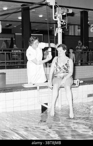 Croix-Rouge aides bénévoles à Sportfondsbad [pour] personnes handicapées Hôpital Croix-Rouge B'wijk Croix-Rouge, Whizgle News, Dutch Desk, pays-Bas, 1950 - 2000 on 30-11-1978. Ces rubriques apparaissent dans l'image. La scène se déroule dans une zone de piscine, caractérisée par une atmosphère stérile et clinique. L'éclairage est lumineux, éclairant l'espace avec une lueur légèrement dure, soulignant les surfaces carrelées et se reflétant sur l'eau. Au premier plan, une femme est assise sur un rebord peu profond, les pieds immergés dans l'eau. Elle est habillée d'un maillot de bain à motifs aux formes géométriques audacieuses Banque D'Images