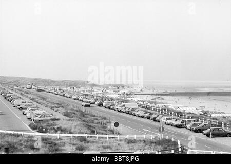 Parking à IJmuiden plage plage et plages IJmuiden pays-Bas, Whizgle News, Dutch Desk, pays-Bas, 1950 - 2000 on 04-08-1982. Ces rubriques sont illustrées dans l'image. La scène capture un large cadre côtier où une longue étendue de plage est visible au loin, encadrée par de douces dunes et un horizon clair. Le premier plan présente une chaussée bordée de véhicules stationnés, créant une rangée organisée qui transmet un sentiment d'activité animée. Les voitures sont principalement dans des tons discrets, tels que le gris, le noir et le blanc, correspondant à l'esthétique monochrome globale. Sur la plage, il y a un SC Banque D'Images