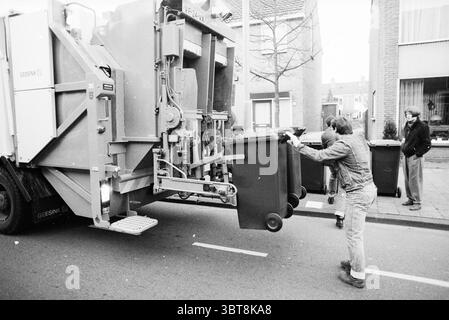 Collecte des seaux à ordures, Whizgle News, Dutch Desk, pays-Bas, 1950 - 2000 sur 19-11-1989. Ce sont les éléments de l'image. Dans un cadre urbain animé, une scène se déroule le long d’une rue résidentielle. Un grand véhicule de collecte des déchets domine la vue, sa carrosserie métallique peinte dans des tons de gris. Le véhicule est immobile, accompagné d'une légère brume d'échappement, ce qui laisse entendre son caractère opérationnel. Au premier plan, une personne vêtue d'une tenue décontractée avec une veste distincte se plie légèrement pour manœuvrer une poubelle de couleur foncée vers l'arrière du camion. Les roues du bac sont visibles, sli Banque D'Images