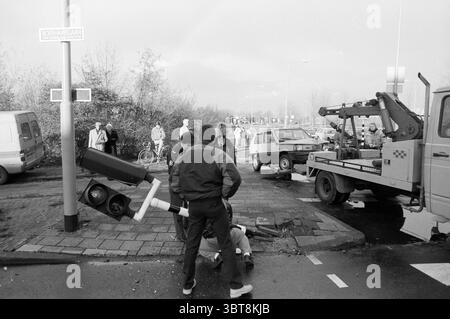 Dommages à un feu de circulation après un accident de voiture Haarlem Boerhaavelaan pays-Bas, Whizgle News, Dutch Desk, pays-Bas, 1950 - 2000 on 11-1988. L'image contient ces rubriques. Dans une scène urbaine animée, un moment chaotique se déroule où un véhicule a clairement rencontré un accident à une intersection. L'accent est mis sur plusieurs personnes qui sont rassemblées autour, leurs expressions révélant l'inquiétude et l'engagement avec ce qui se passe. Un groupe de passants, y compris une personne portant un manteau foncé, se tient sur le côté pour observer la situation. D'autres sont directement impliqués ; un individu appea Banque D'Images