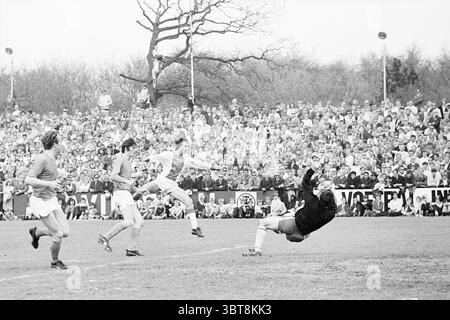 Match de football Telstar-Ajax dans IJmuiden IJmuiden pays-Bas, Whizgle News, Dutch Desk, pays-Bas, 1950 - 2000 sur 10-05-1970. Ces rubriques sont illustrées dans l'image. Dans la scène, un moment dramatique se déroule sur un terrain herbeux, probablement un terrain de sport. L'accent est mis sur un joueur qui saute dans les airs, se préparant à frapper une balle avec une expression intense. Il porte un maillot lumineux qui contraste avec les tons terreux du terrain, parsemé de taches d'herbe verte. A proximité, deux défenseurs sont positionnés stratégiquement, l'un accroupi bas et l'autre debout, tous deux portant u Banque D'Images