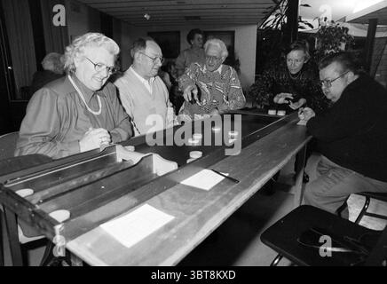 Ouverture de la maison Arnolduspark Hoofddorp Arnolduspark pays-Bas, Whizgle News, Dutch Desk, pays-Bas, 1950 - 2000 le 20-12-1991. Ce sont les éléments de l'image. Dans un cadre intérieur confortable, un groupe de personnes âgées se réunit autour d'une longue table en bois qui domine la scène. La table est sombre avec une surface brillante, reflétant la lumière ambiante au-dessus. Sur la table, de petits objets circulaires sont disposés, probablement partie d'un jeu dans lequel ils sont engagés. Assis à la table sont cinq personnes, chacune affichant des expressions variées de concentration et d'amusement. A gauche, une femme avec Banque D'Images