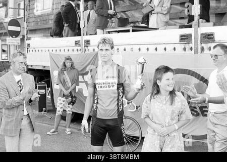 Tour de Zwanenburg - cérémonie des gagnants Zwanenburg, Whizgle News, Dutch Desk, pays-Bas, 1950 - 2000 le 23-07-1990. L'image inclut ces rubriques. Au premier plan, un cycliste se tient fièrement debout, tenant un trophée brillant en hauteur avec un regard de triomphe sur son visage. Il est habillé dans un kit de cyclisme vibrant, principalement rouge et noir, qui contraste avec les tons discrets de la scène environnante. La texture de sa tenue suggère qu'elle a été conçue pour la performance, épousant étroitement sa monture. À côté de lui, une fille se tient avec les cheveux touffus, portant une robe florale légère qui ajoute une touche de bri Banque D'Images