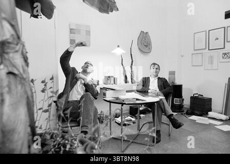 Artiste Sjoerd Buisman et Servaas M. de Nijs, Whizgle News, Dutch Desk, pays-Bas, 1950 - 2000 le 07-01-1996. Ces rubriques sont illustrées dans l'image. La scène capture deux hommes assis dans un espace de travail contemporain et minimaliste. Un homme, à gauche, fait des gestes animés avec sa main gauche levée et sa main droite tenant un petit objet, démontrant peut-être une idée ou un concept. Il a une expression réfléchie, des lunettes perchées sur son nez, et est habillé d'une veste sombre sur une chemise de couleur claire. L'autre homme, à droite, a une posture plus détendue, assis avec confiance avec un smi léger Banque D'Images