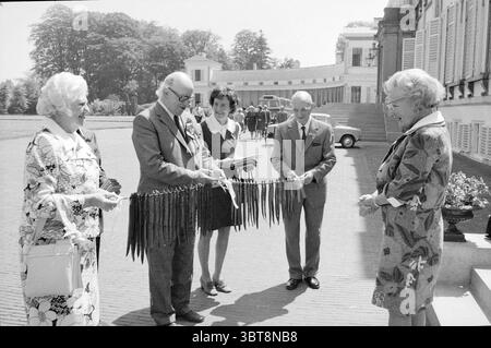 Personnes âgées visitant la reine Juliana au Palais Soestdijk Soestdijk, Whizgle News, Dutch Desk, pays-Bas, 1950 - 2000 on 05-06-1974. Voici les rubriques de l'image. Dans un cadre extérieur ensoleillé, un groupe de cinq personnes est réuni, créant une scène ciblée et engageante. La douce lumière illumine leurs visages, soulignant la chaleur et la camaraderie entre eux. Deux femmes se tiennent à gauche, toutes deux élégamment vêtues, avec une dans une robe à motifs qui présente de grands motifs floraux audacieux dans des teintes principalement blanches et sombres. L'autre femme, portant une tenue plus subtile, a un e joyeux Banque D'Images