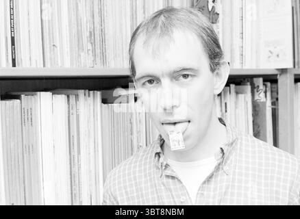 Joost Swarte Cartoonist dessins, Whizgle News, Dutch Desk, pays-Bas, 1950 - 2000 le 14-08-1985. L'image inclut ces rubriques. La scène met en scène un jeune homme avec une expression réfléchie, debout sur fond de nombreux livres ou magazines soigneusement arrangés. Ses cheveux sont courts et de couleur claire, et son visage porte un soupçon de désagrément. Il porte une chemise à carreaux, qui ajoute une touche de chaleur décontractée à son apparence, avec des couleurs telles que le rouge et le bleu en avant dans le motif. Dans un geste ludique, il a un petit morceau de papier qui dépasse de sa bouche, créant une intrigue Banque D'Images