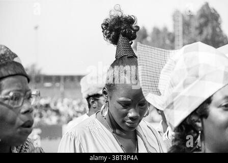 Haarlem - Robin Hood Football Haarlem, Whizgle News, Dutch Desk, pays-Bas, 1950 - 2000 le 15-08-1976. Ce sont les éléments de l'image. La scène capture un moment rempli d’émotion et d’expression culturelle. Au premier plan, une femme se tient debout, la tête légèrement courbée, mettant en valeur une coiffure frappante qui présente une haute torsion en forme de cône. Sa peau scintille doucement sous la lumière du soleil, suggérant une journée chaude. Elle porte de grandes boucles d'oreilles cerceau qui attrapent la lumière, ajoutant une touche d'élégance à son apparence. Autour d'elle sont d'autres individus, chacun exposant des styles uniques et ex Banque D'Images