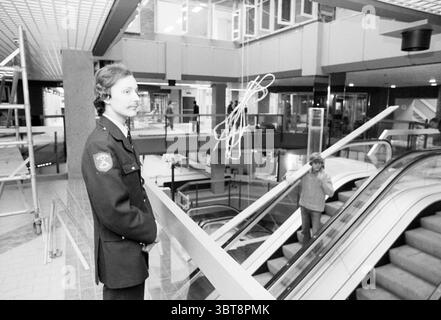 Service de sécurité au complexe Brinkmann Brinkman passage Haarlem Grote Markt pays-Bas, Whizgle News, Dutch Desk, pays-Bas, 1950 - 2000 on 03-03-1982. Voici les rubriques de l'image. La scène se déroule dans un environnement intérieur spacieux et moderne, caractérisé par un design architectural épuré et minimaliste. La structure comporte plusieurs niveaux, avec un atrium ouvert important qui permet à la lumière naturelle de filtrer, bien que l'ambiance générale soit quelque peu atténuée, suggérant une journée couverte à l'extérieur. Au premier plan, un jeune gardien de sécurité se tient confiant, vêtu d'un uniforme sombre que Banque D'Images